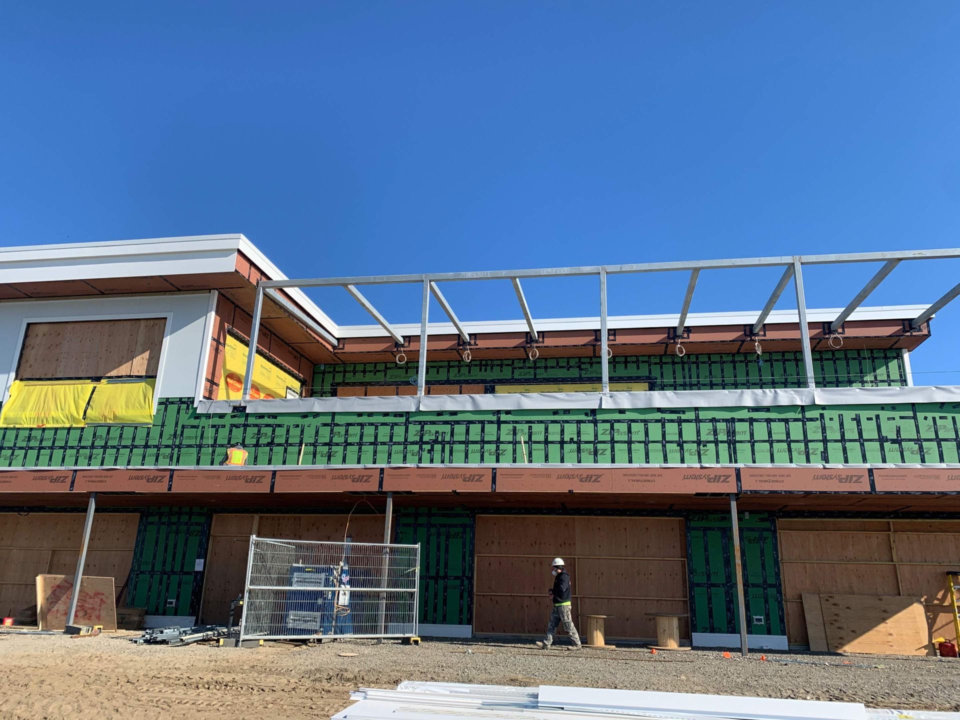 Construction site with building frame, insulation, and a worker walking. Blue sky overhead.