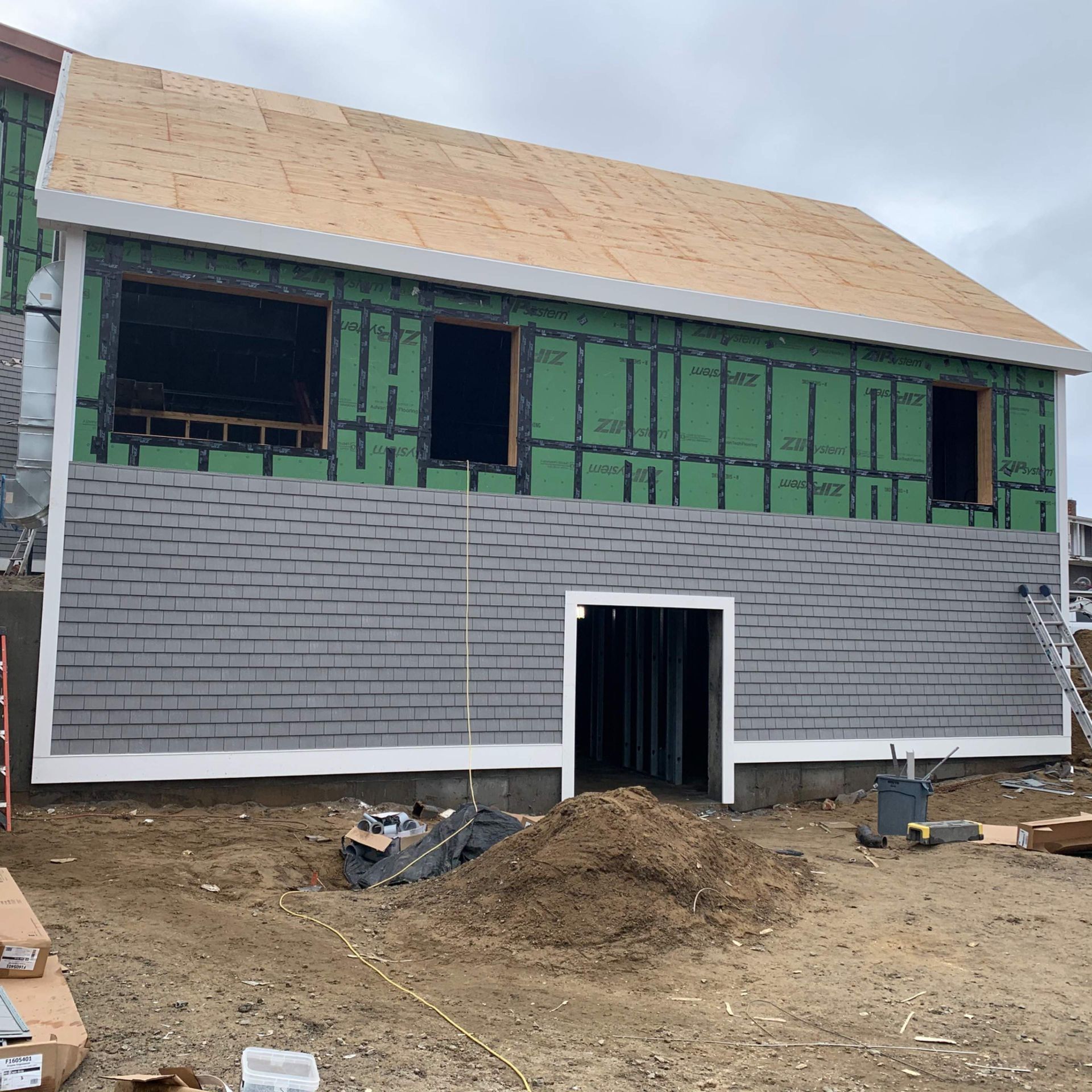 Two-story building under construction with gray shingle siding, green sheathing, and plywood roof.