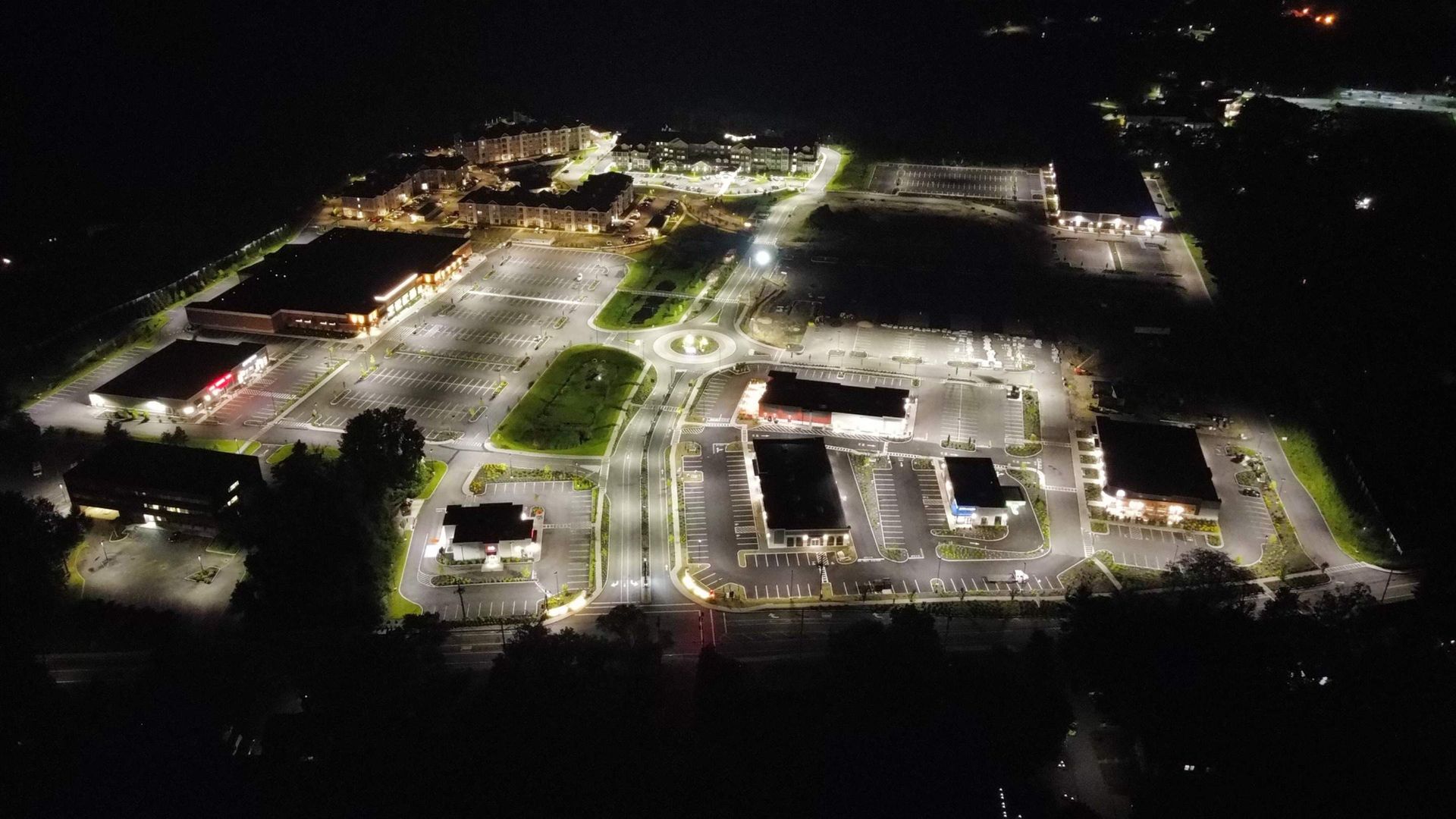 Nighttime aerial view of a brightly lit shopping center and parking lots.