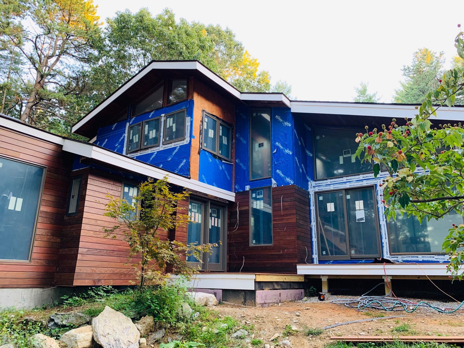 Modern two-story house under construction, with wooden siding, blue wrap, and large windows.