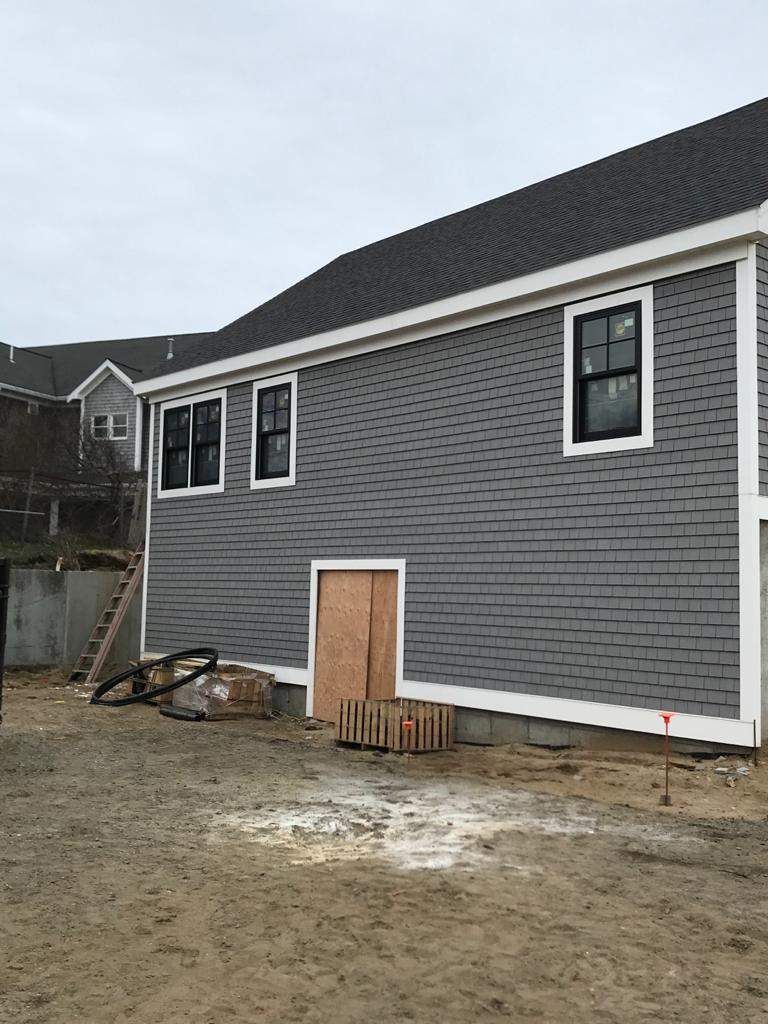 Gray-shingled house with black-framed windows, a wood door, and a gravel yard; construction in progress.