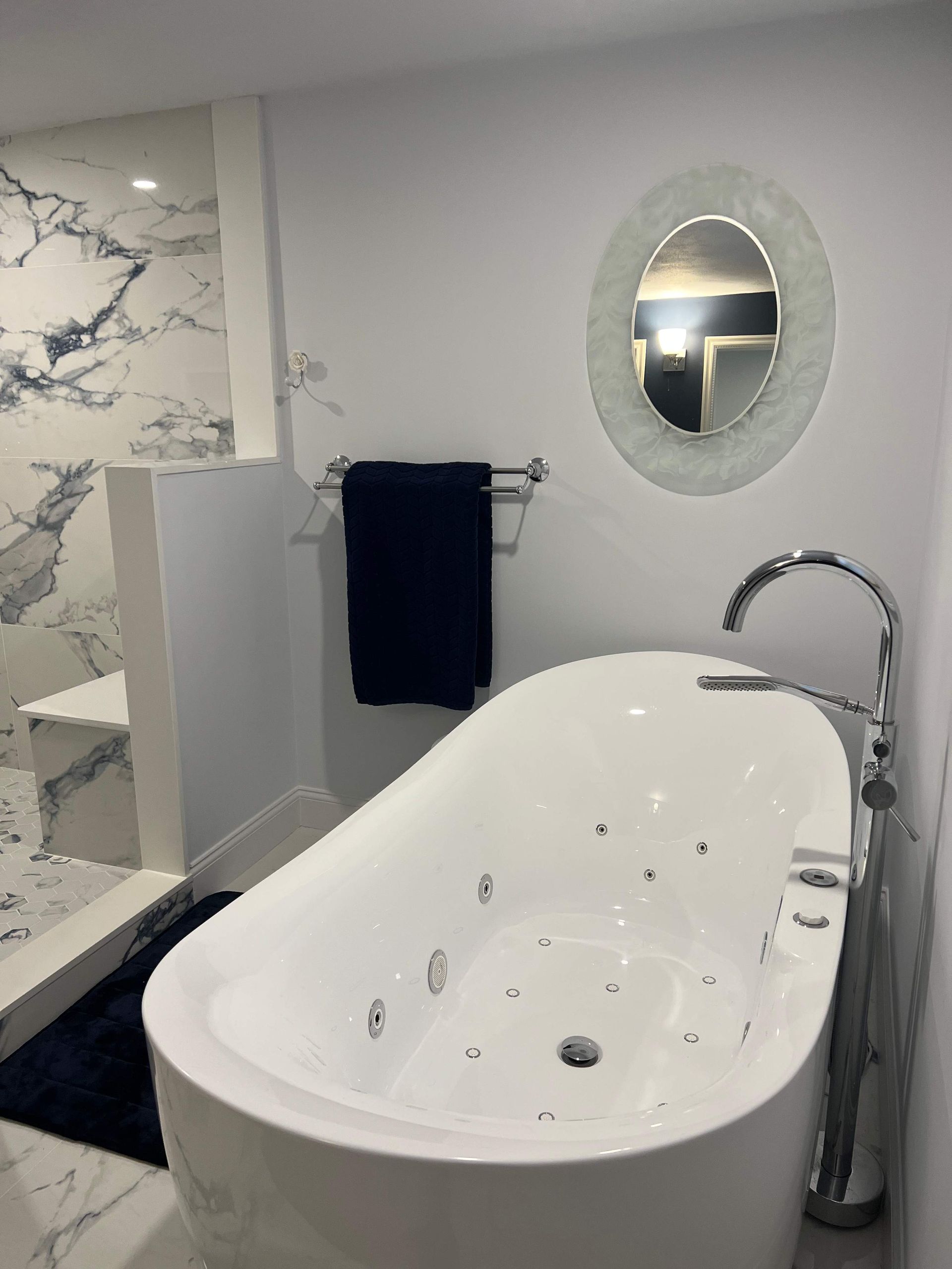 White bathtub with chrome fixtures in a modern bathroom with marble walls and a round mirror.