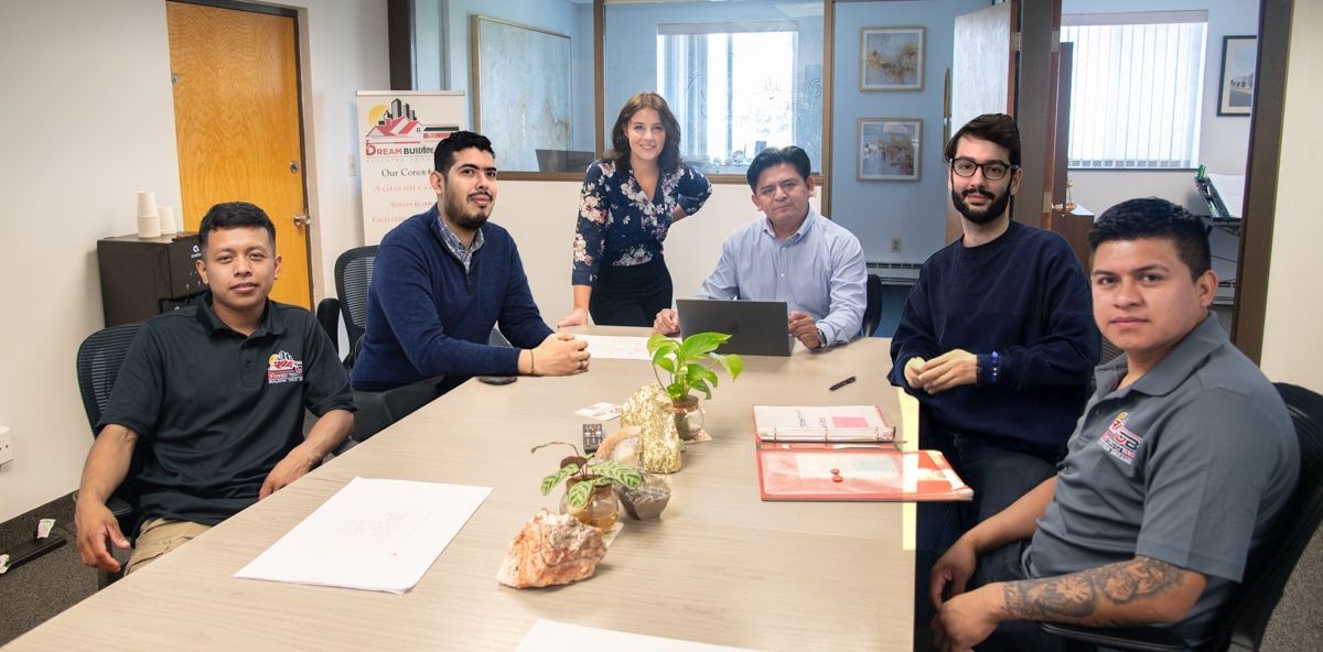 Seven people seated around a table in an office. They are looking at the camera. A laptop and plant are on the table.