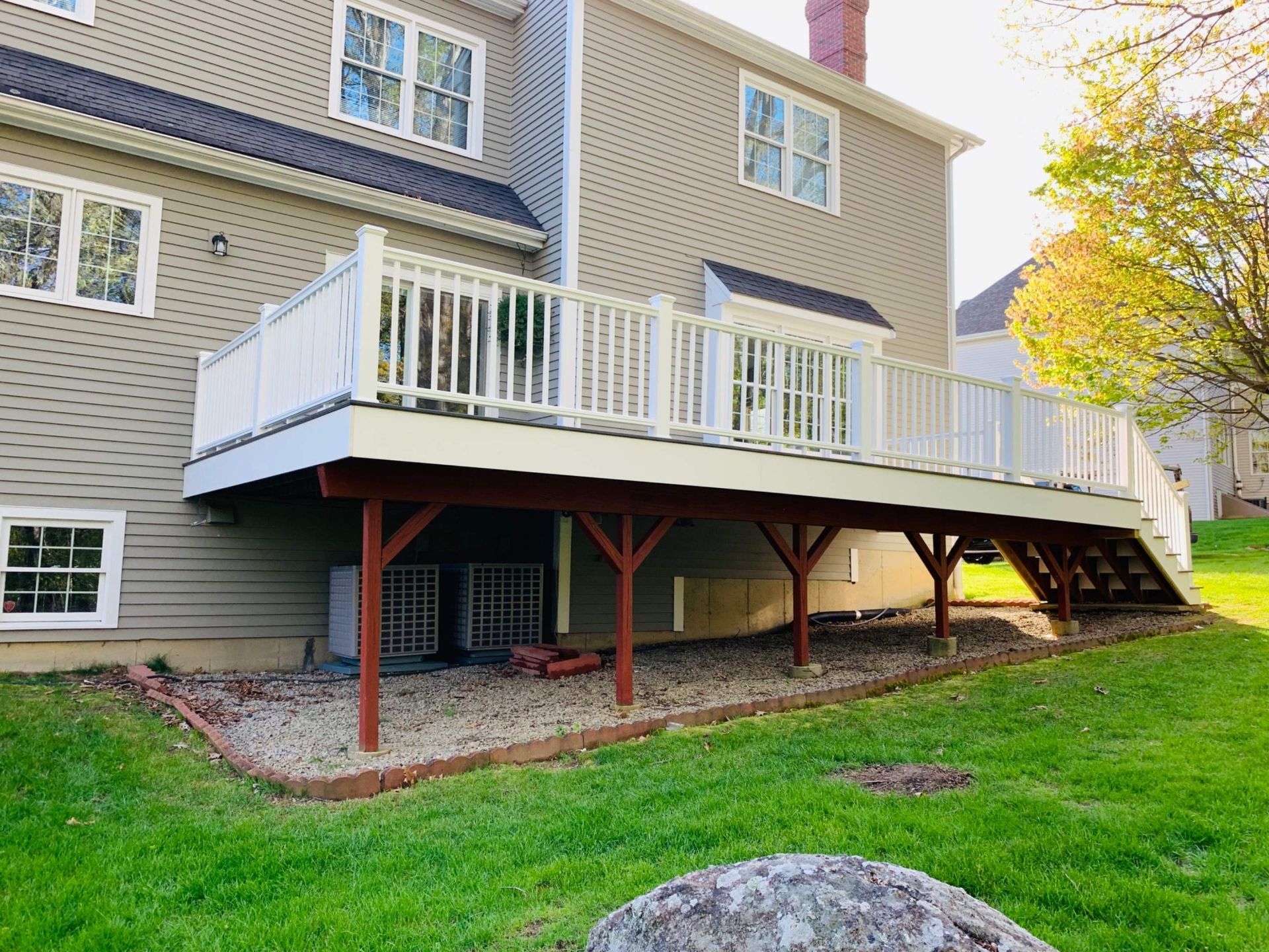 Back of a beige house with a white deck, wooden support beams, and steps to the ground.