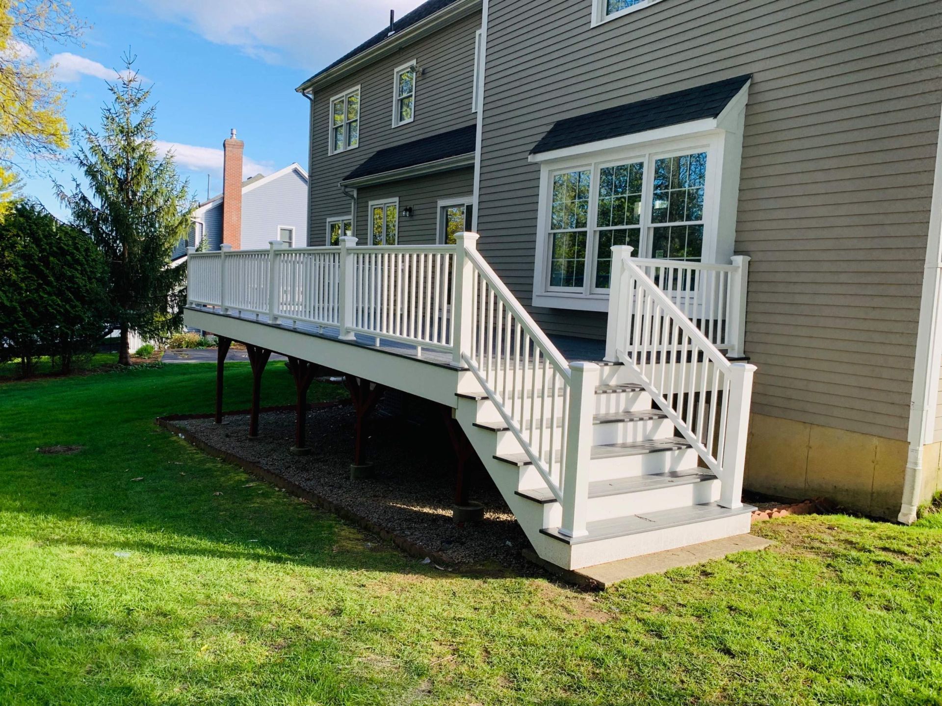 White wooden deck with stairs attached to a two-story gray house on a green lawn.