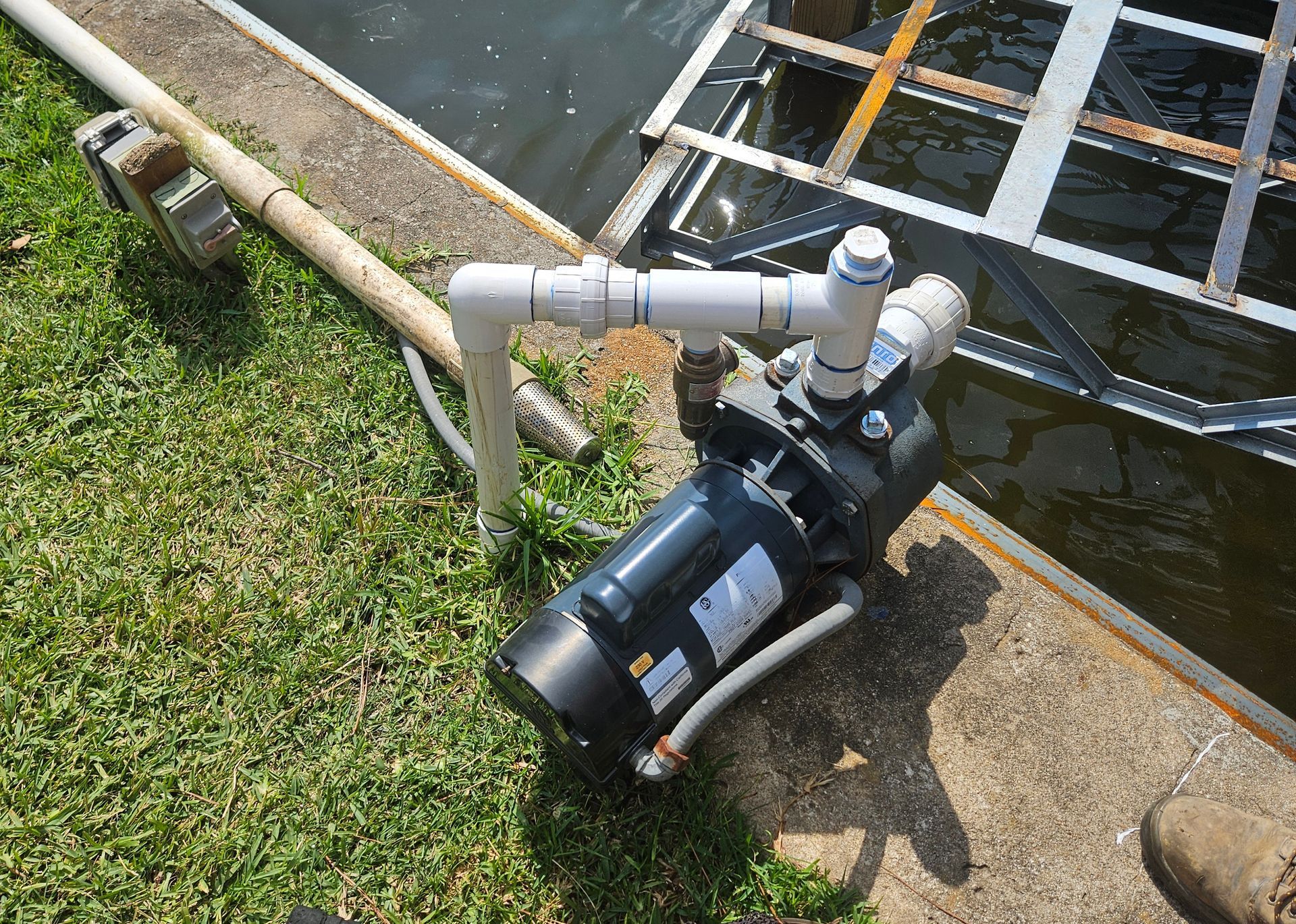 Water pump with white PVC pipes on grass near a body of water and metal structure.