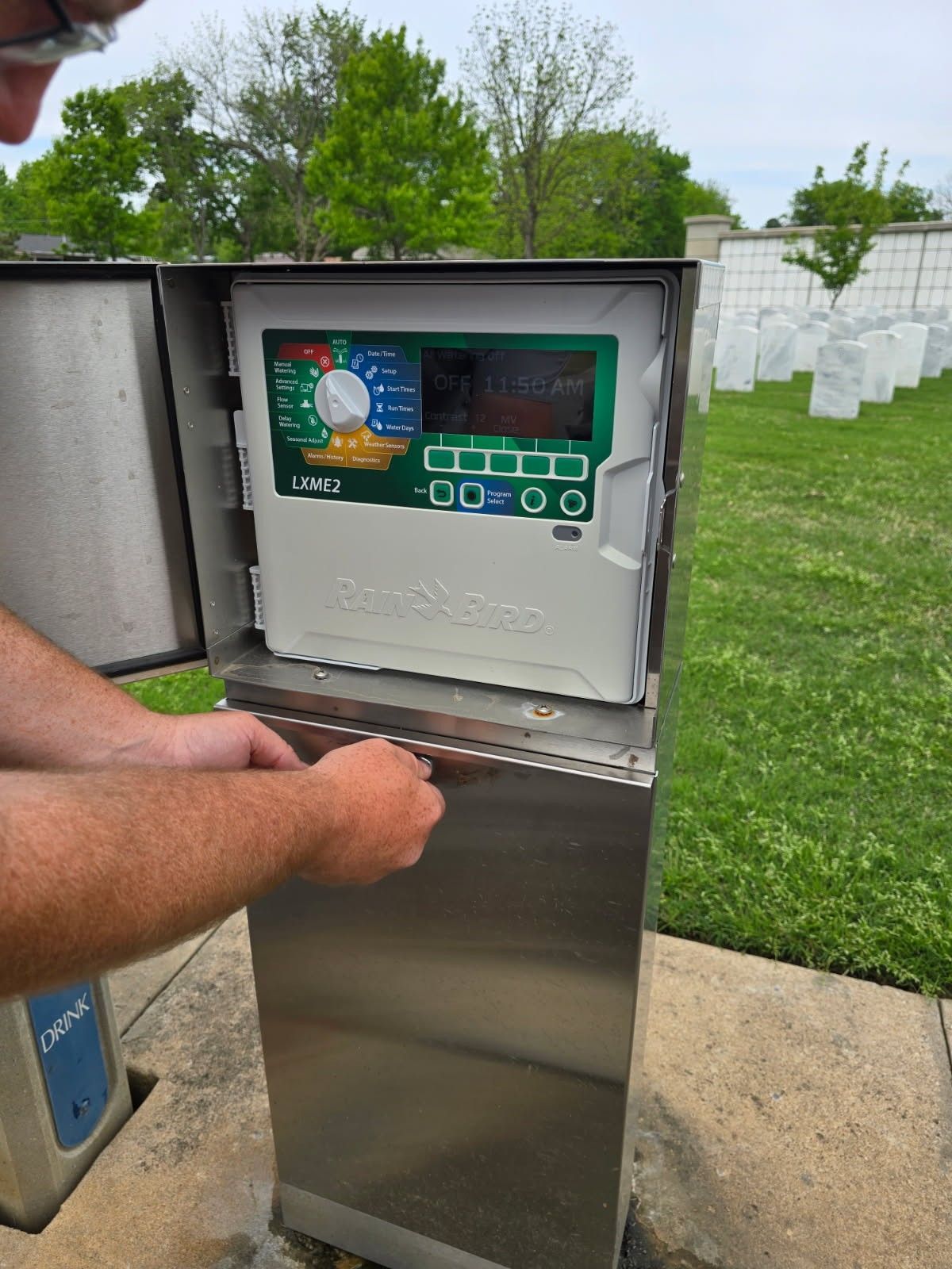 Person opening a stainless steel irrigation control box outdoors near headstones and trees.