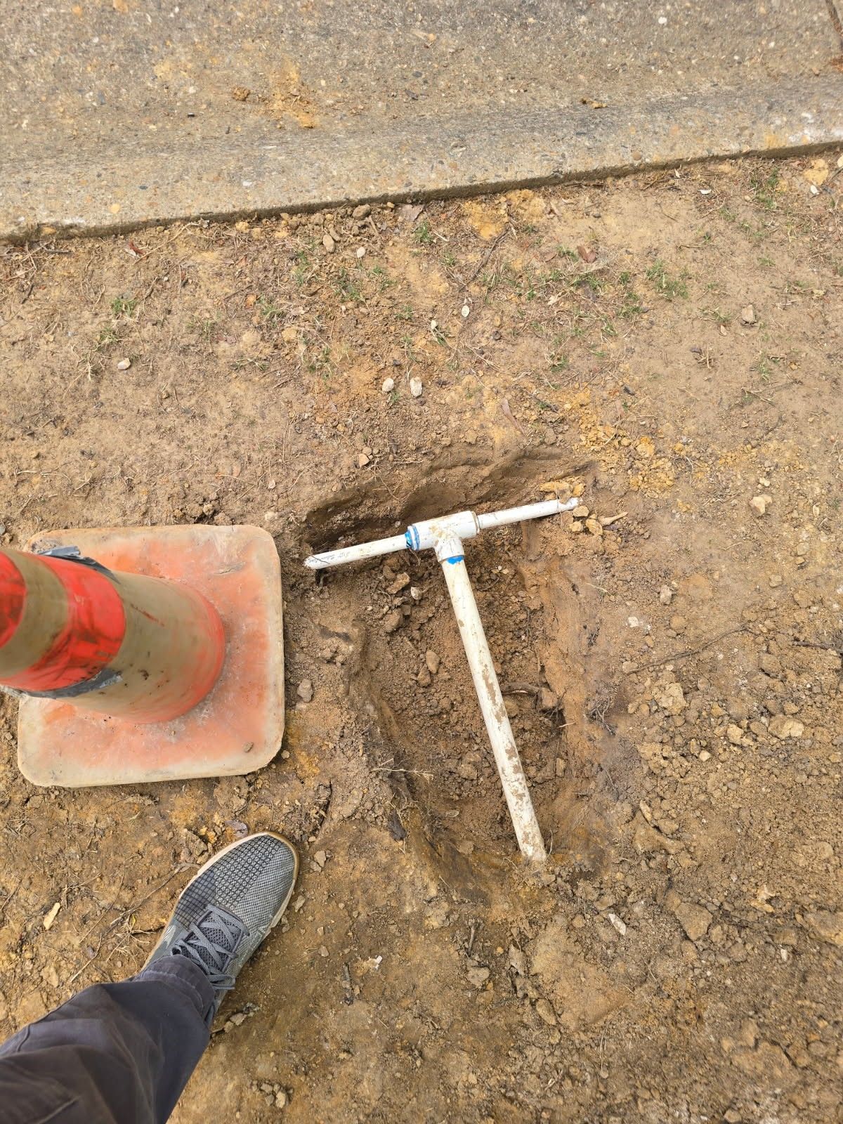 PVC pipe in dirt, near a cone and sidewalk. A foot is also visible.