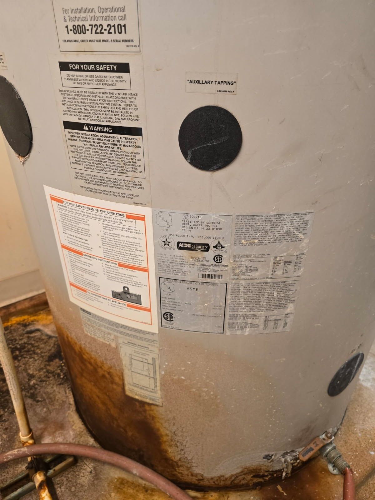 Leaking water heater with rust and labels. The floor is stained with water.