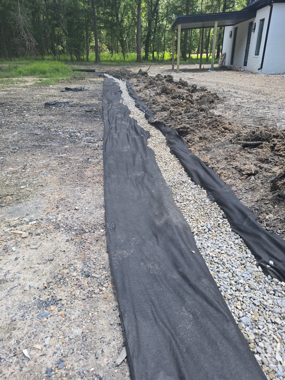A black fabric-lined trench with gravel and dirt, next to a gravel driveway and a house with a porch in a wooded setting.