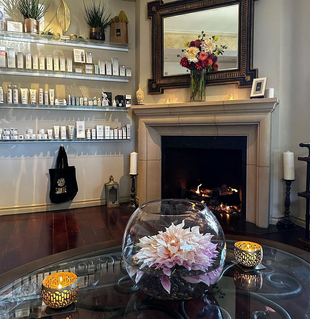 A cozy room features a fireplace with a mirror and flowers above, shelves of products, and a glass table with a centerpiece.