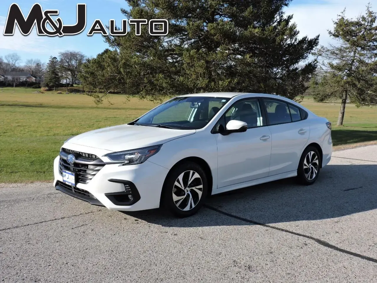 White sedan parked on a roadside with grass and trees in the background, M&J Auto logo in corner