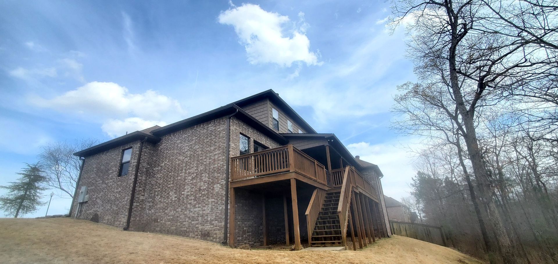A large brick house with a wooden deck is sitting on top of a hill surrounded by trees.