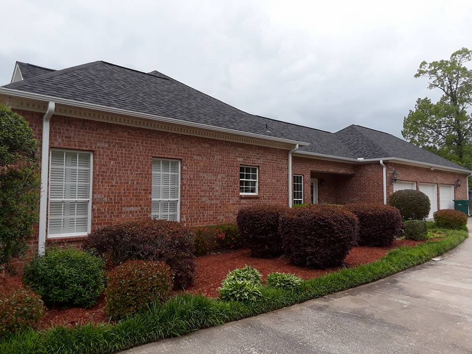 A large brick house with a black roof is surrounded by bushes and shrubs.