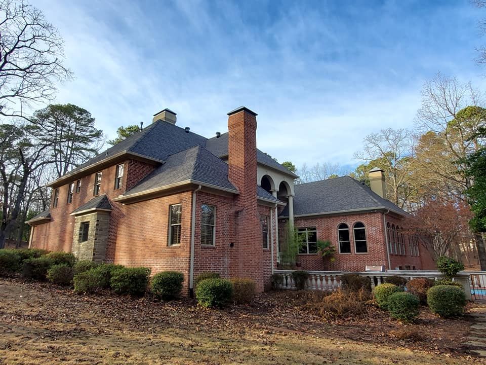 A large brick house with a gray roof is surrounded by bushes and trees.