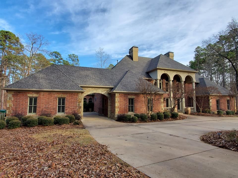 A large brick house with a gray roof is surrounded by trees and leaves.