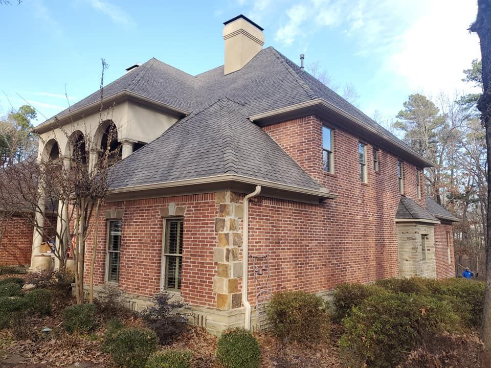 A large brick house with a gray roof is surrounded by bushes and trees.