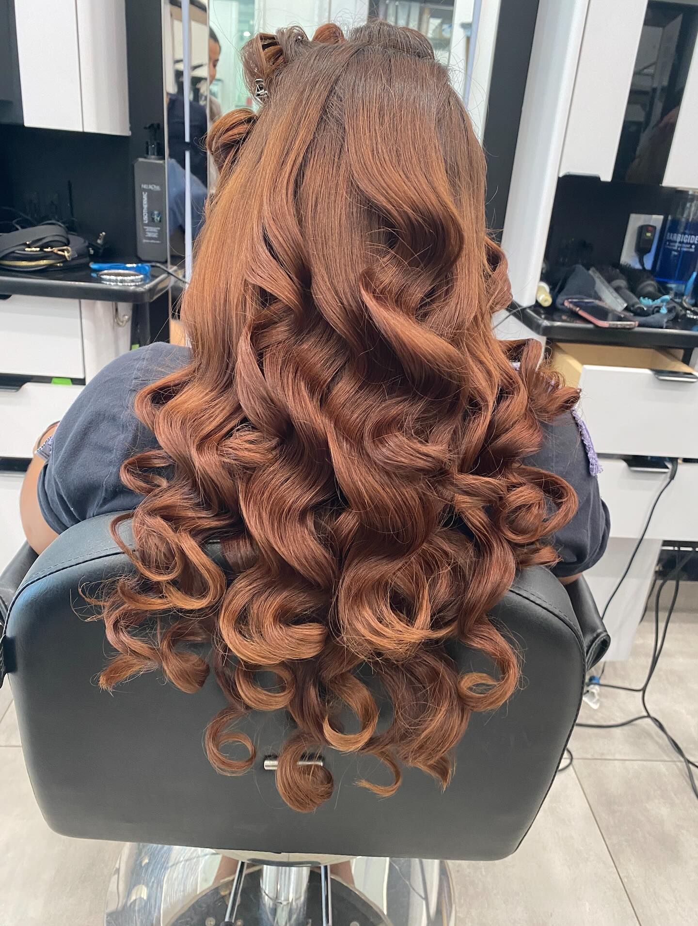 Long curly auburn hair styled half-up, cascading down the back of a seated person in a salon chair