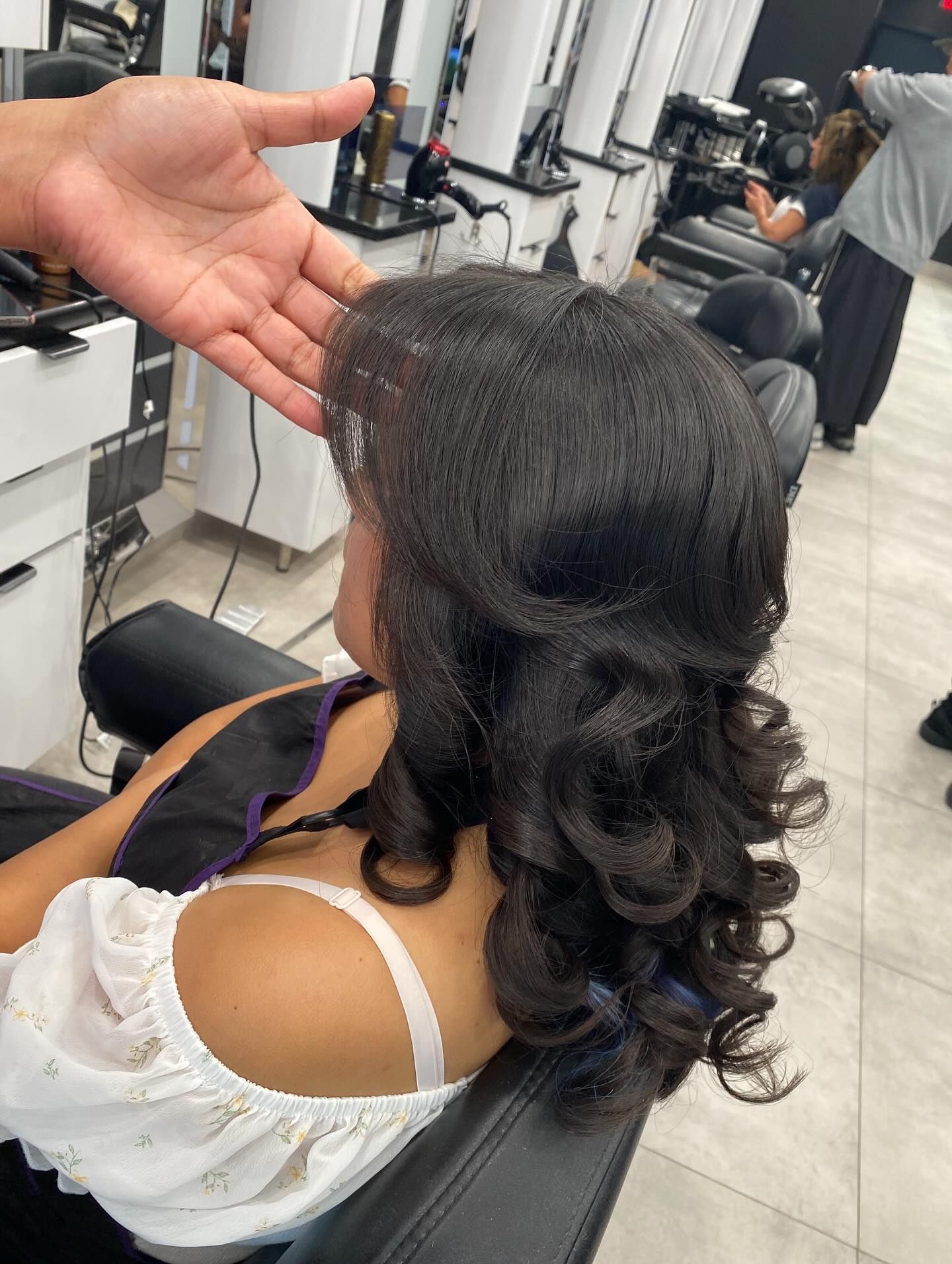 Woman with long dark curls in a salon chair, shown from behind while a stylist checks her hair.