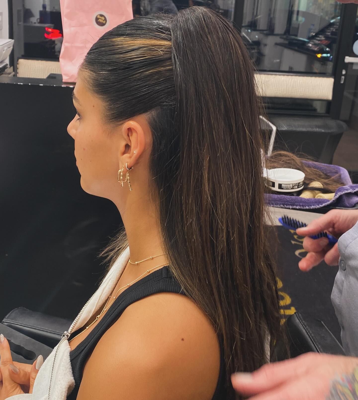 Woman with a high ponytail and braided side, seated in a salon while hairstyling tools are in use.