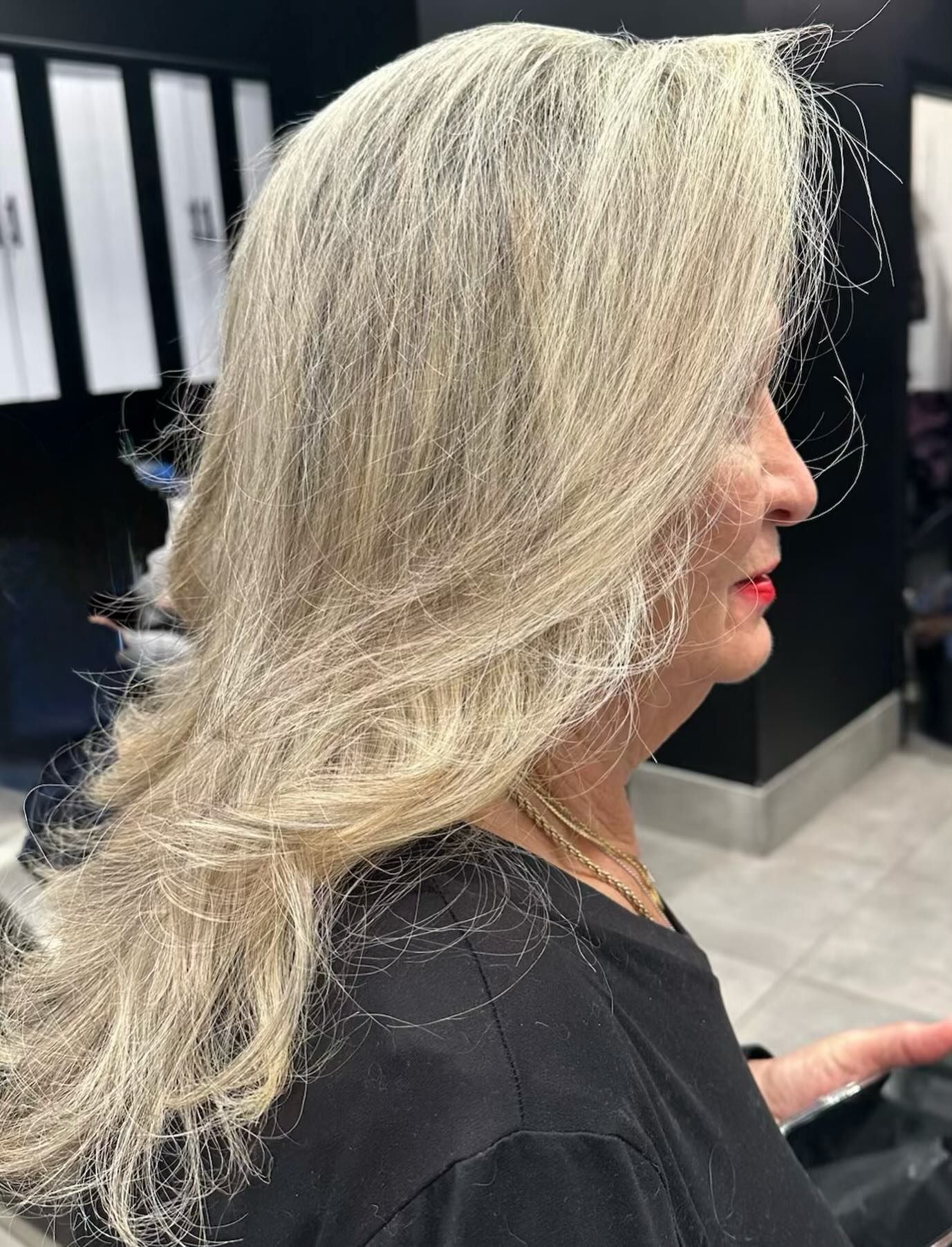 Side profile of a woman with long silver-blonde hair in a salon chair, wearing a black top.