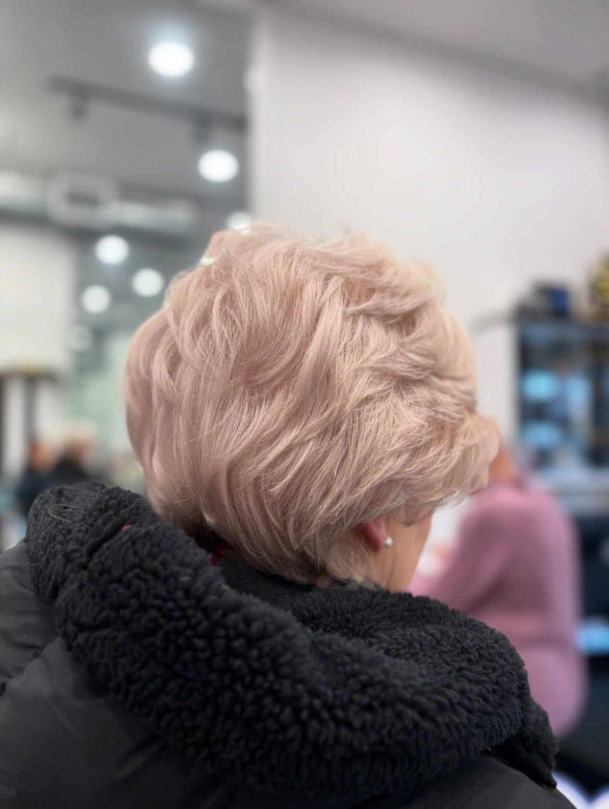 Back view of a person with short pink hair wearing a black textured coat in a salon.