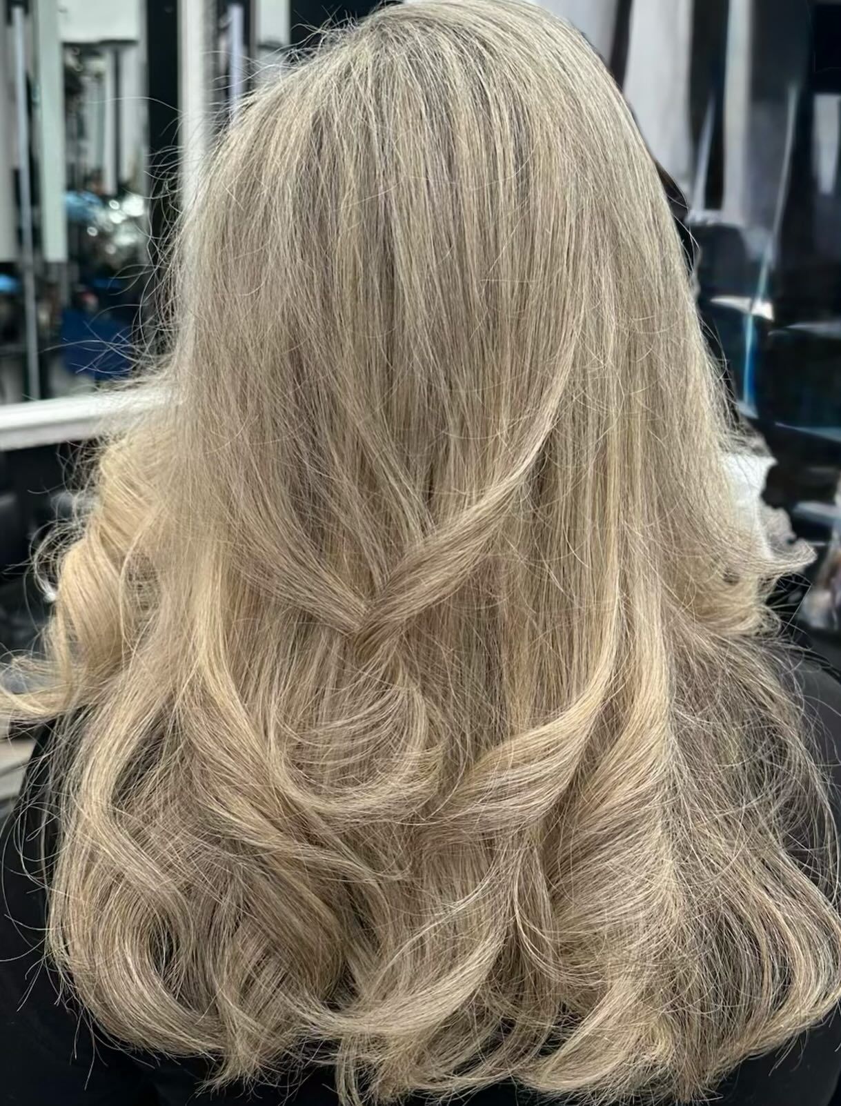 Long layered blonde hairstyle with soft waves, shown from the back in a salon chair.