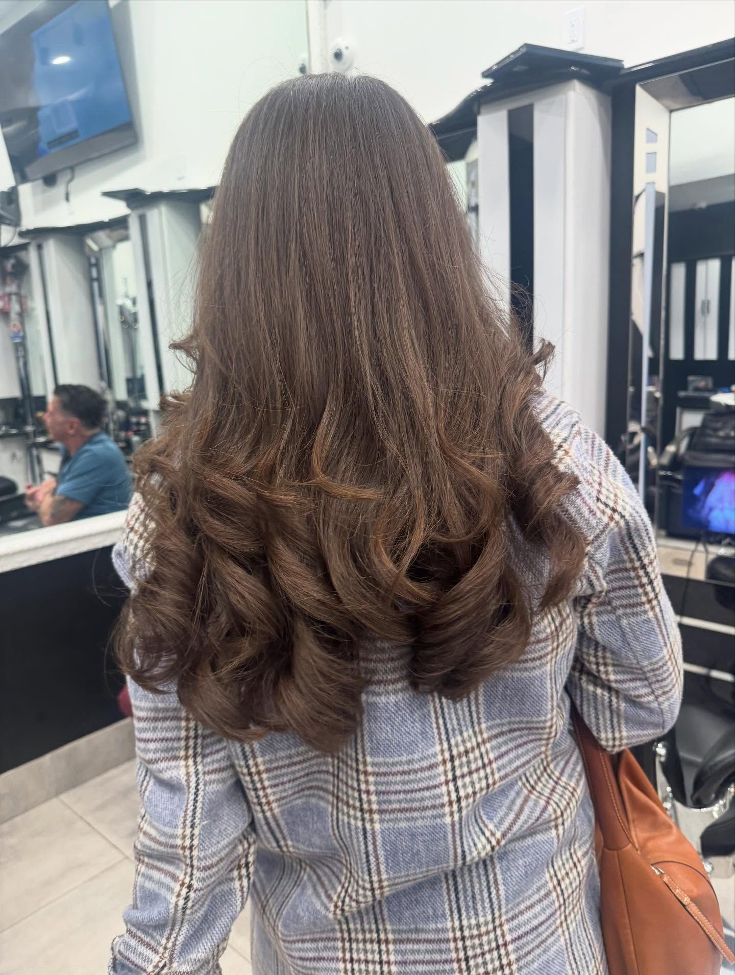 Woman with long curly brown hair seen from behind in a salon, wearing a plaid coat and carrying a brown bag.