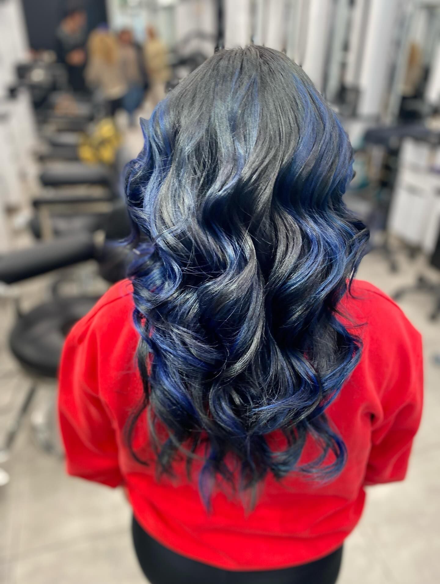 Person with long wavy blue hair wearing a red top in a salon chair, viewed from behind.
