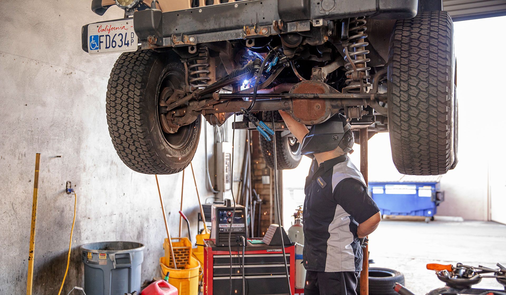 Mechanic inspecting an off-road vehicle lifted on a garage hoist, with tools and equipment below.