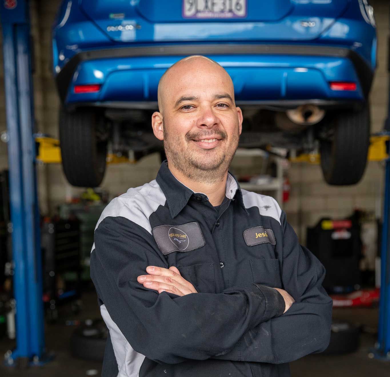 Mechanic standing with arms crossed in a garage, with a blue car raised on a lift behind him