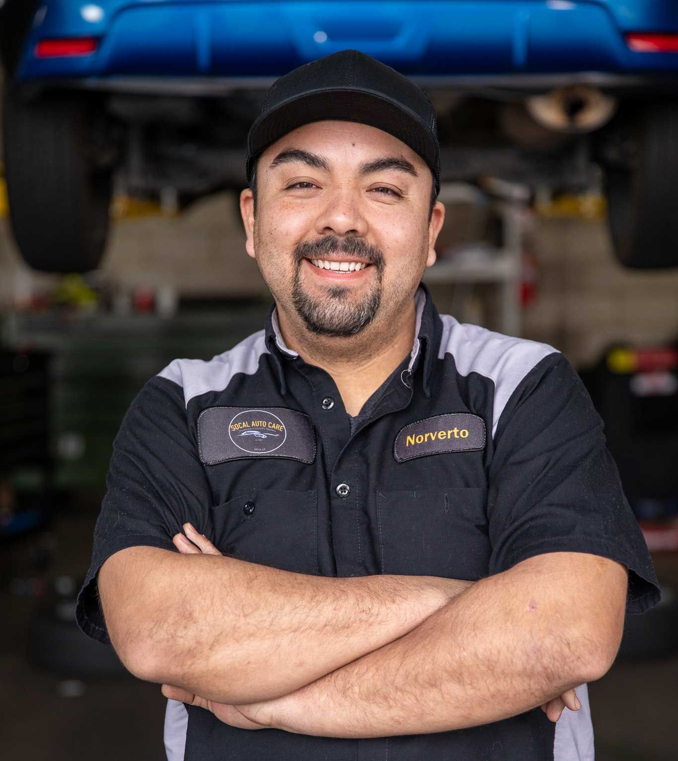 Smiling mechanic with crossed arms standing in a garage beneath a blue car on a lift