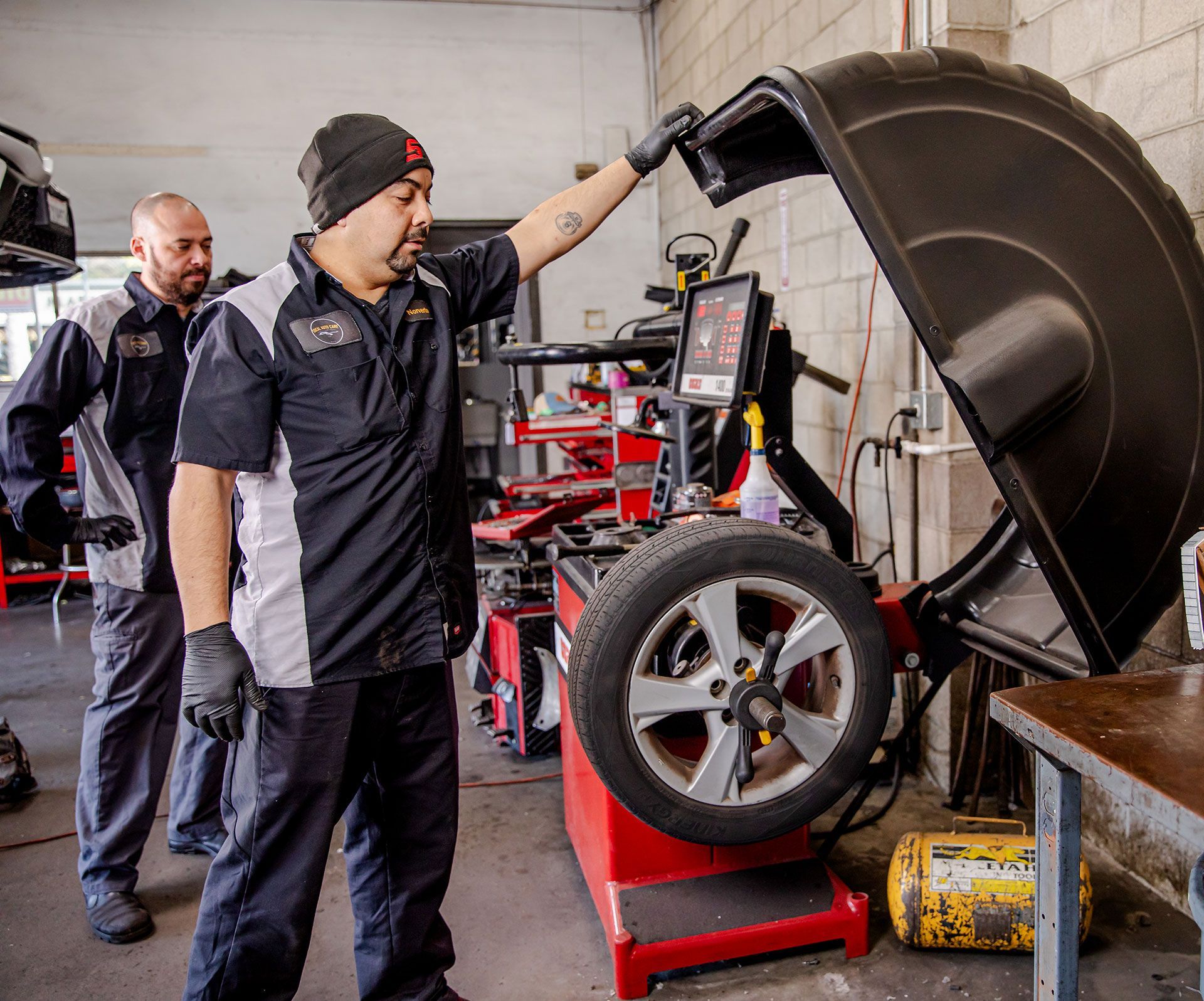 Mechanic in dark uniform lifts car hood in auto shop while another stands nearby.