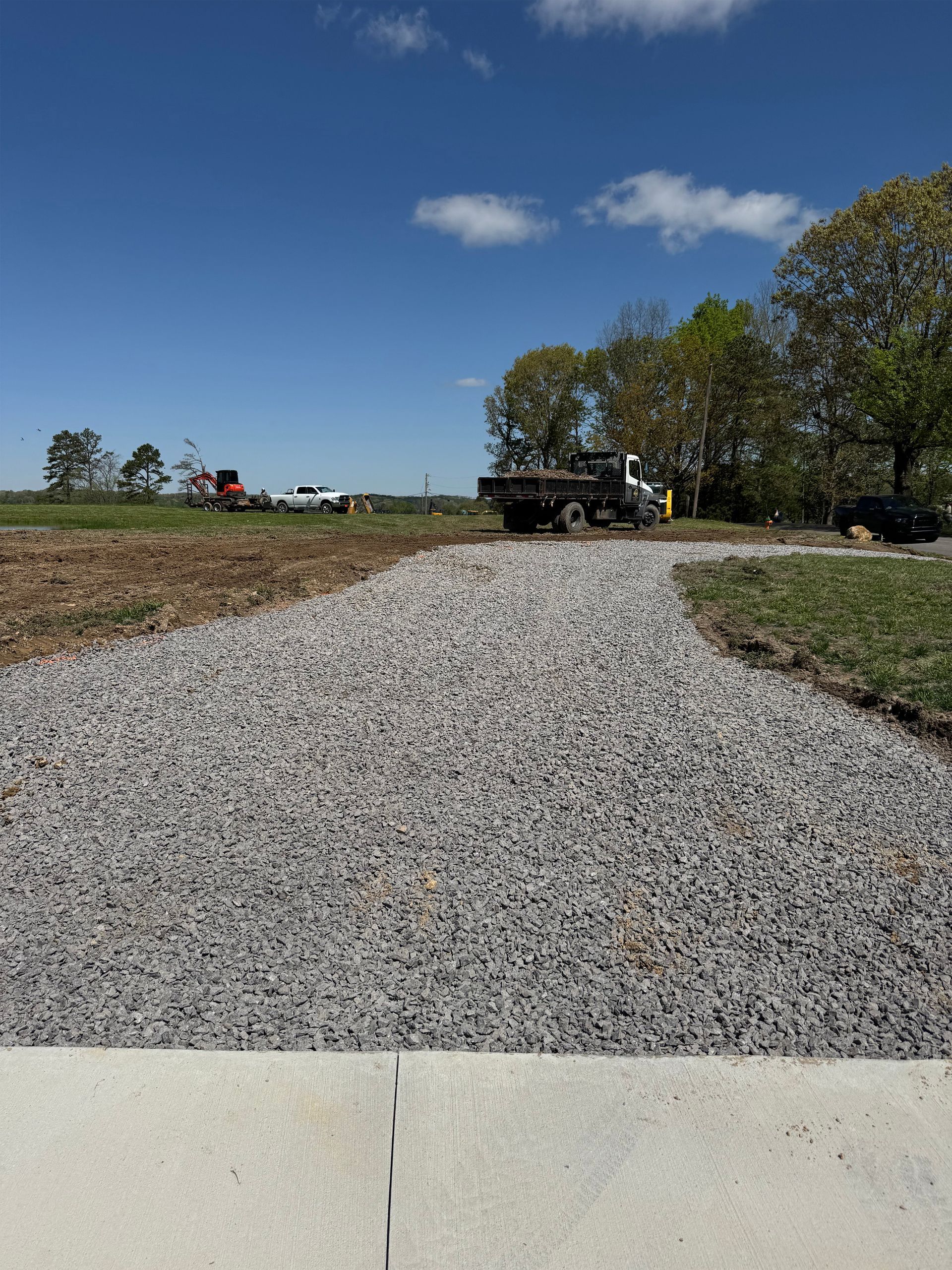 A fresh gravel driveway leads from a concrete edge toward a construction site with a dump truck under a blue sky.