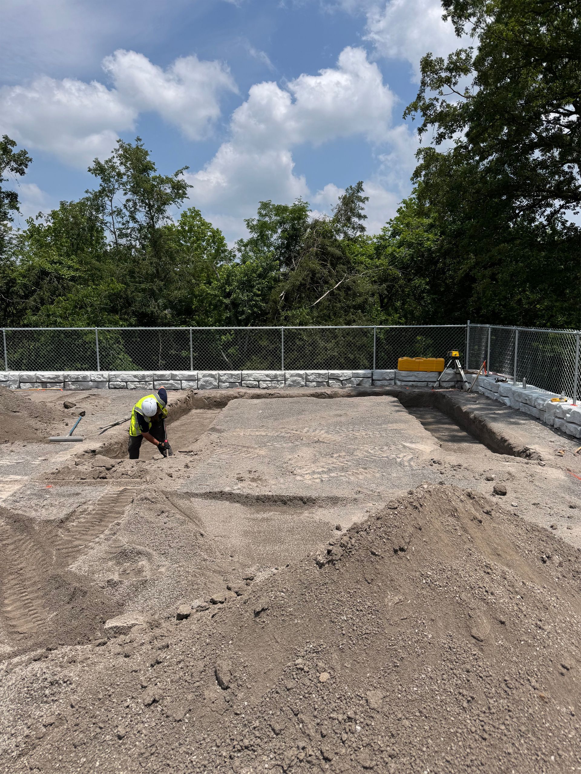 A construction worker in a high-visibility vest digs in a dirt lot enclosed by a chain-link fence under a blue sky.