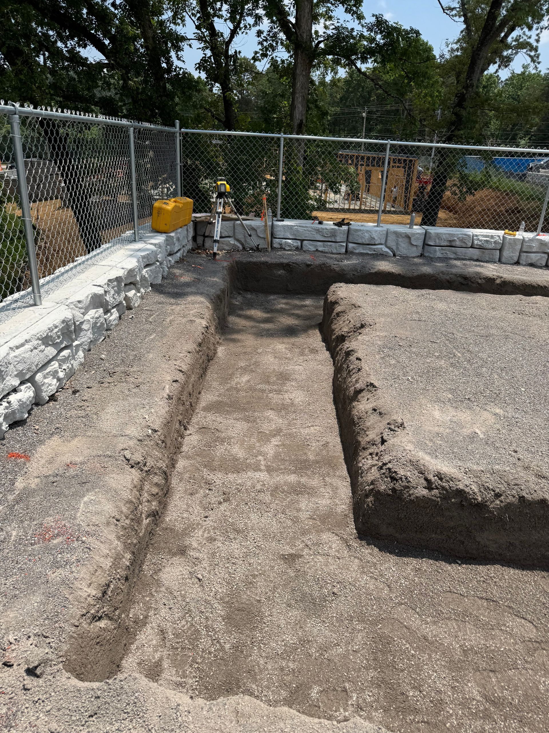 A gravel-filled rectangular construction trench in a yard next to a white retaining wall and chain-link fence.