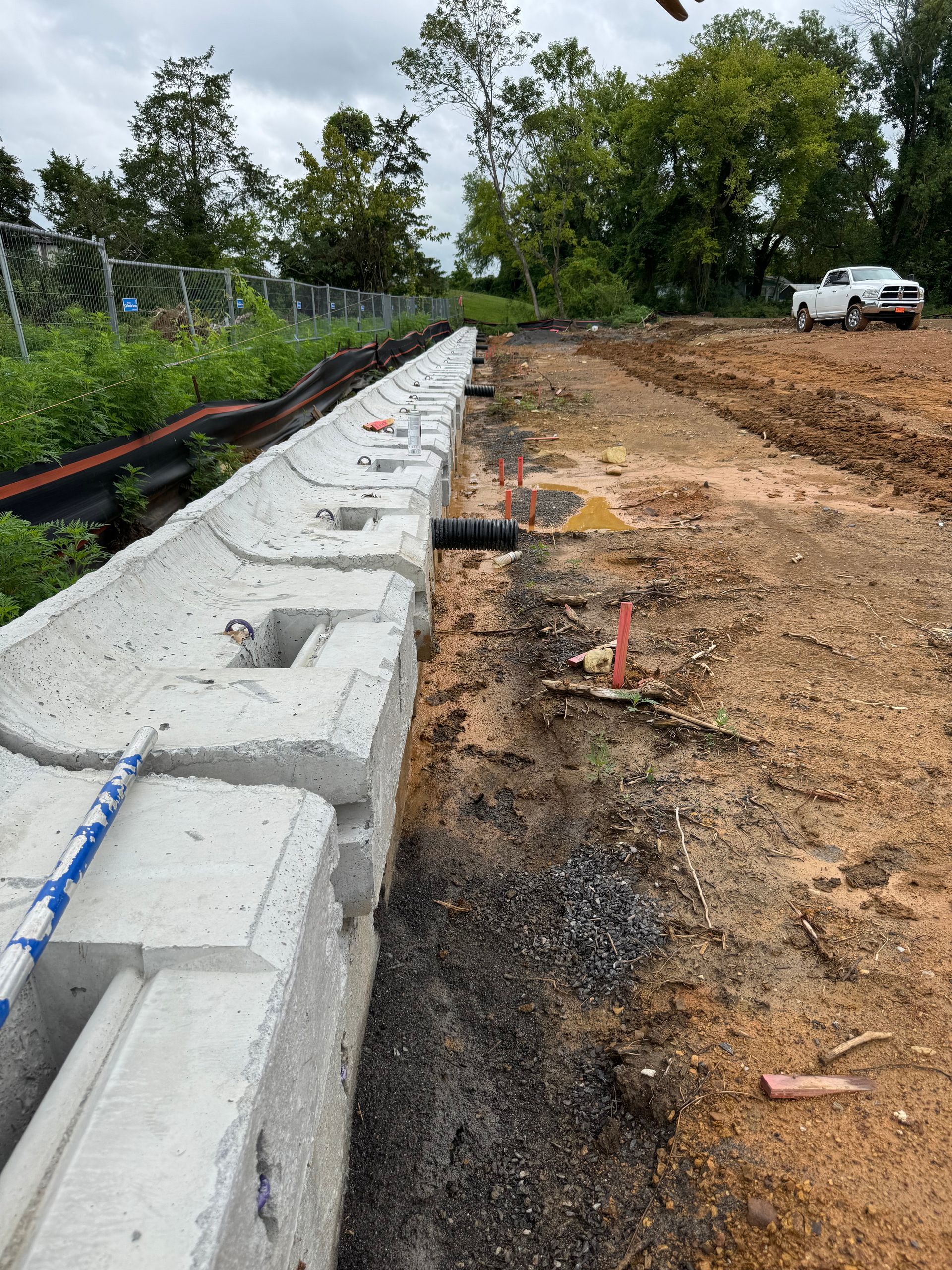 Concrete retaining wall blocks under construction along a dirt embankment with construction stakes and a drainage pipe.
