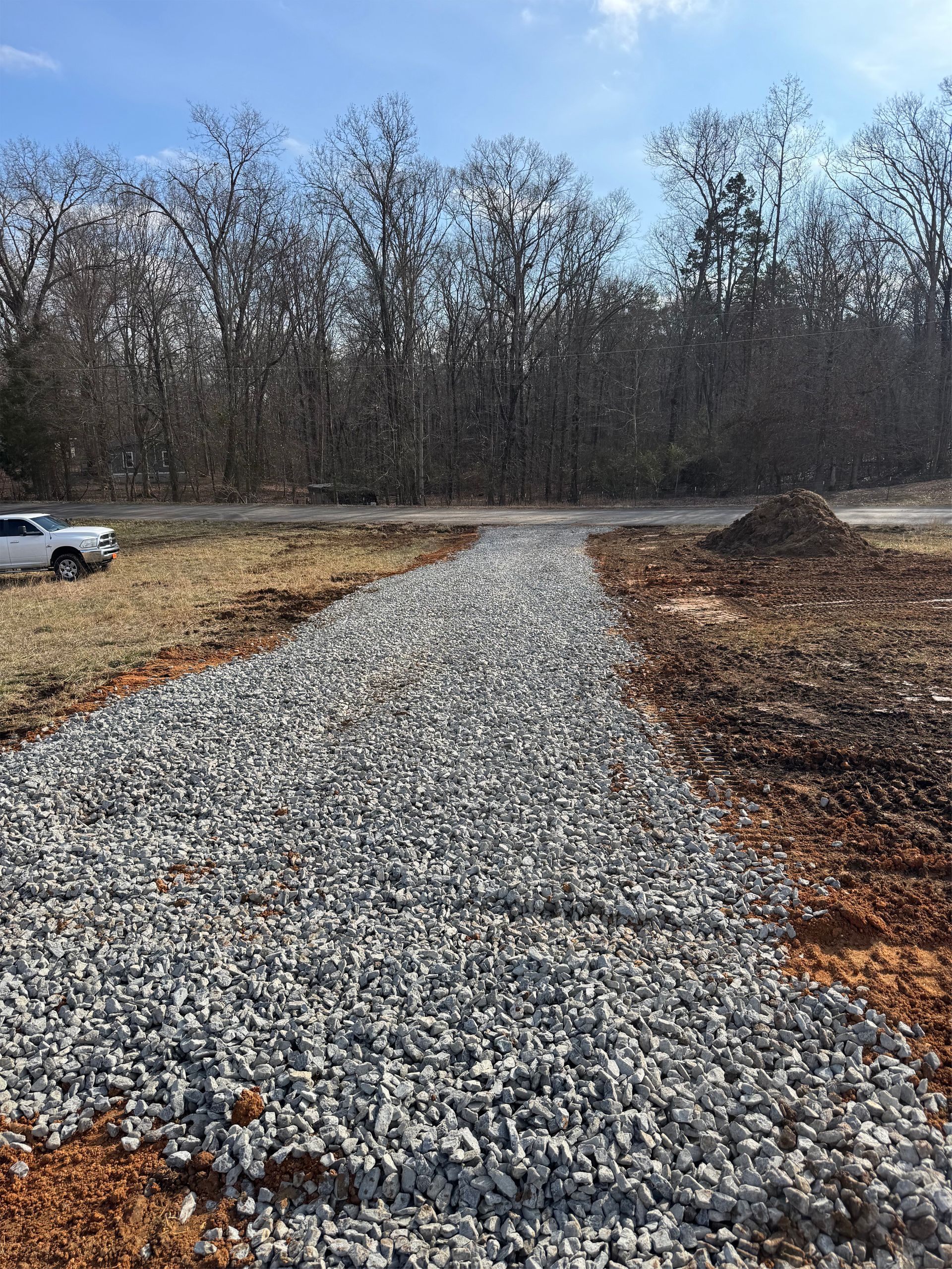 A gravel driveway under construction leads toward a wooded area under a blue sky, with a white truck parked nearby.