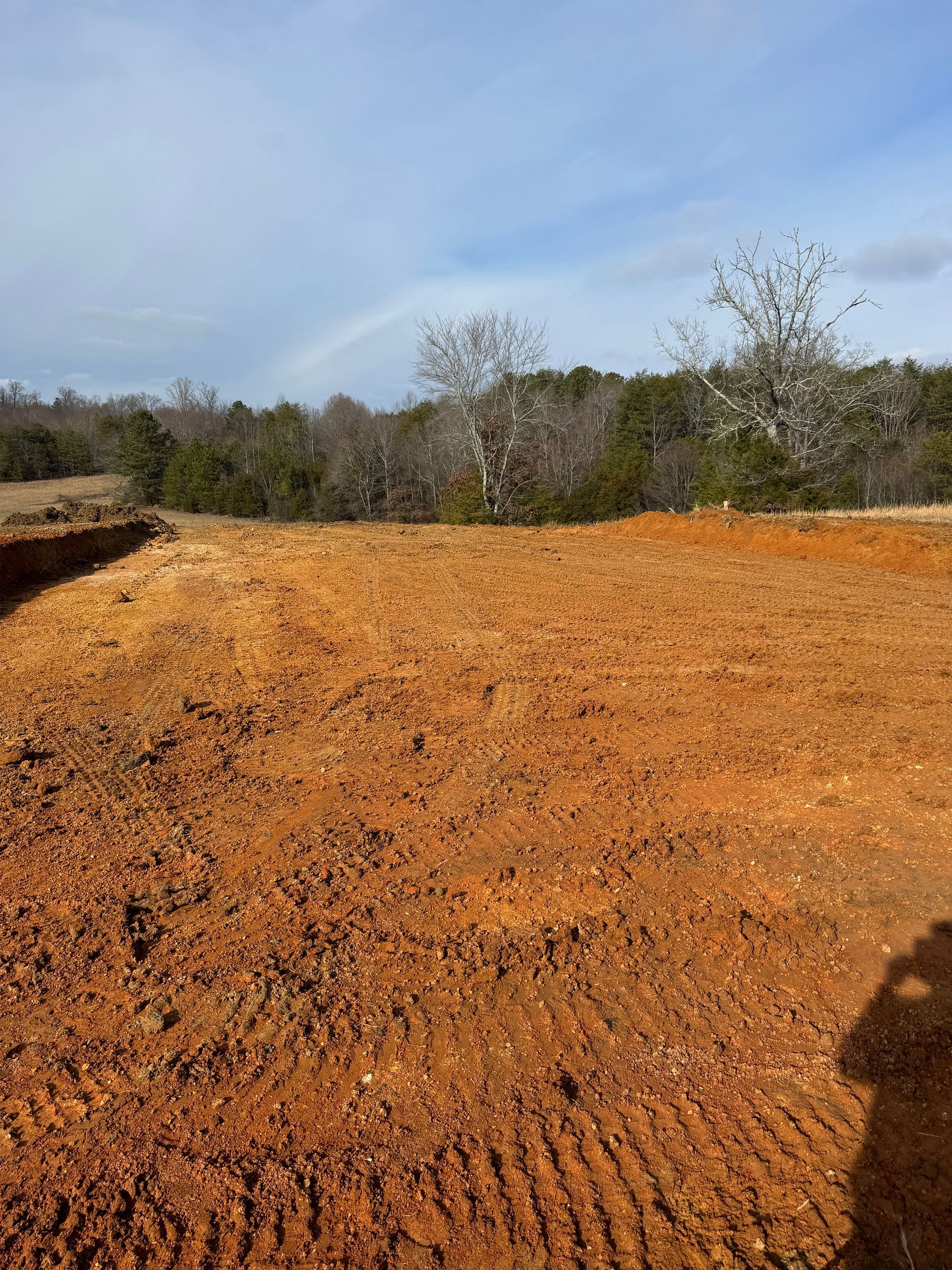 A cleared, orange-clay construction site under a partly cloudy sky, with a line of trees in the background.