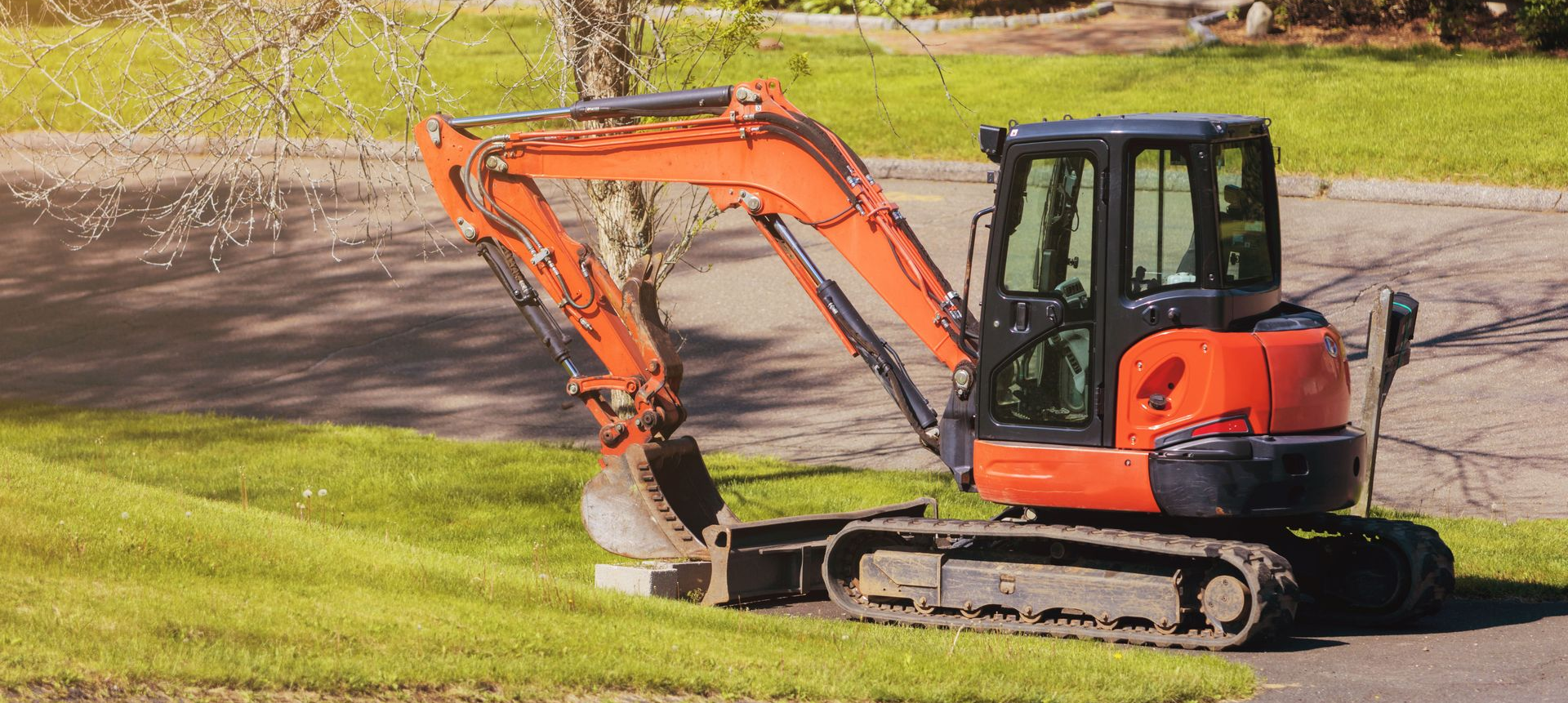 An orange compact excavator parked on a grassy patch next to a paved surface.