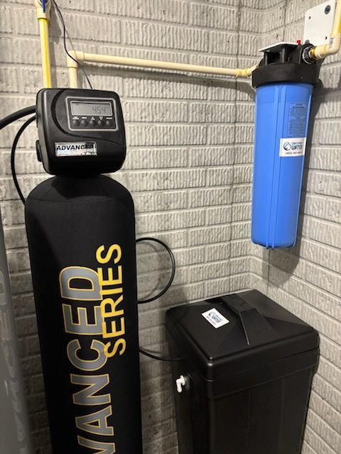 Water softener and filter system installed on a brick wall in a basement. The system includes a black water softener, blue filter, and a salt tank.