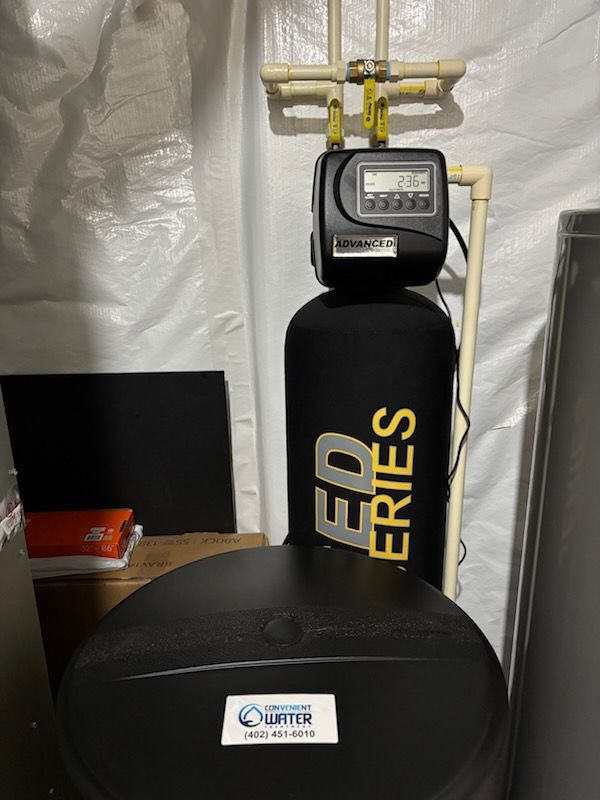 Water softener system in a utility room, featuring a black tank, control unit, and salt storage tank with beige plumbing.