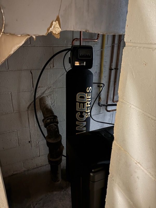 A water filtration system in a basement, composed of a tall black tank with a digital display and connecting pipes.