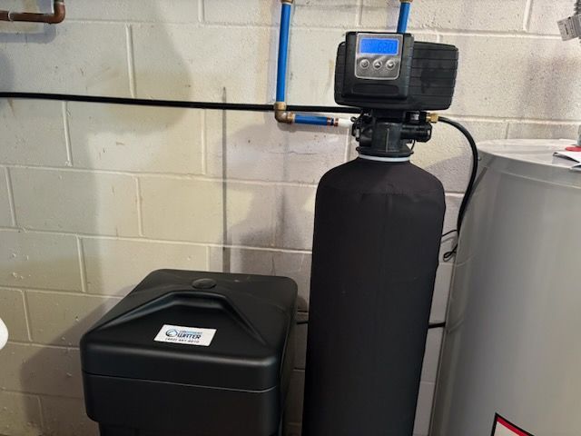 Water softener system installed in a basement, featuring a tall black tank, a black salt storage container, and blue water pipes.