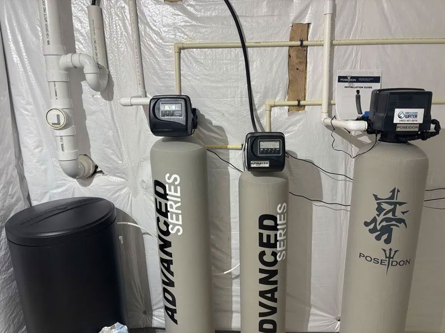 Water filtration system in a utility room; three vertical tanks, pipes, digital displays, and a black trash can.