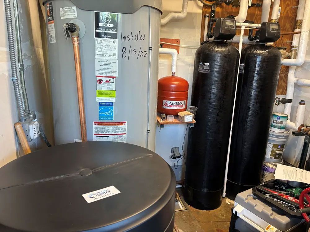 Water heater and filtration system in a basement. Tanks and pipes are visible with tools on the floor.