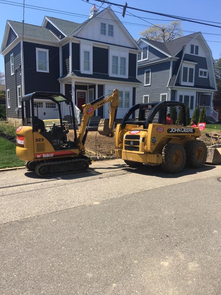 Two construction vehicles are parked on the side of the road in front of a house.