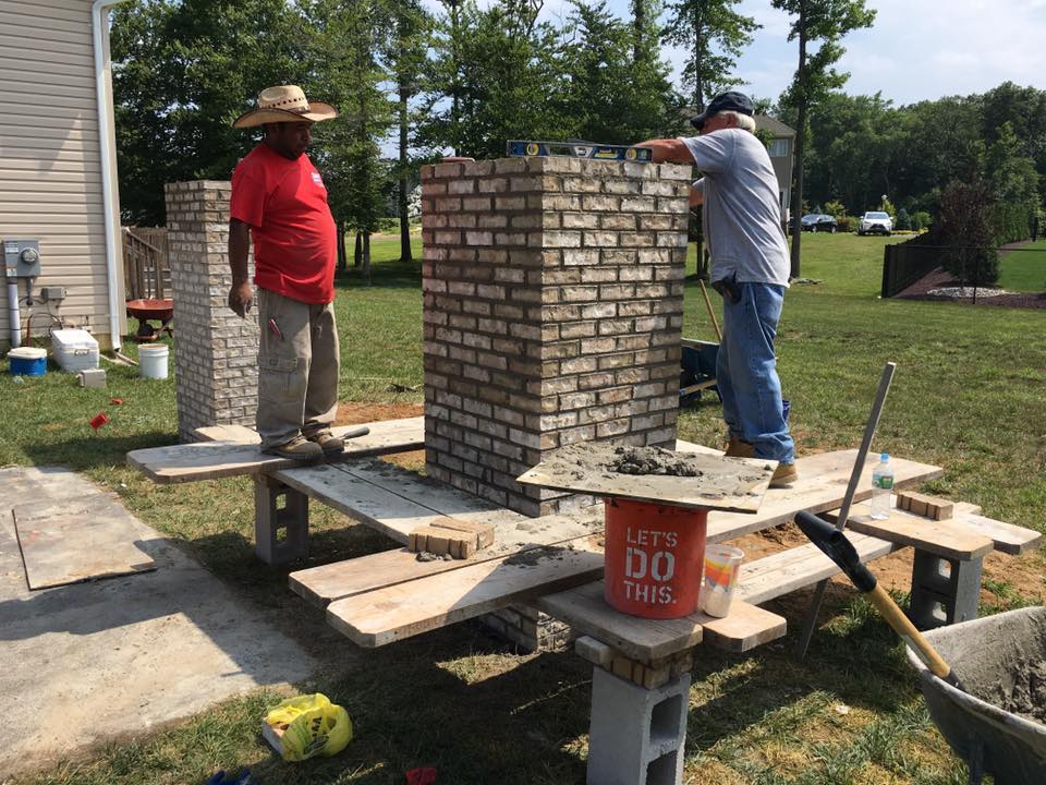 Two men are working on a brick chimney in a backyard.