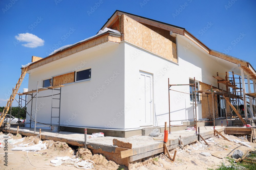 House under construction with white stucco, wooden framing, and scaffolding against a blue sky.