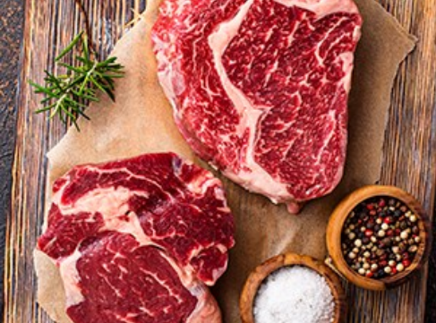 Two raw marbled steaks on parchment with rosemary, salt, and peppercorns on a wooden board